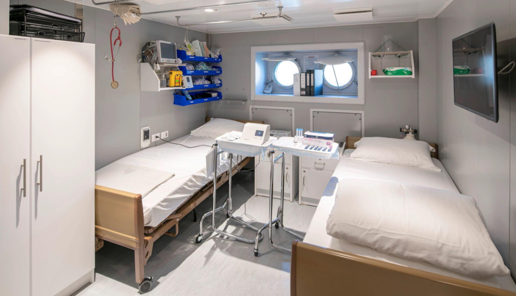 A small medical infirmary on a ship with two hospital beds, medical equipment, and portholes