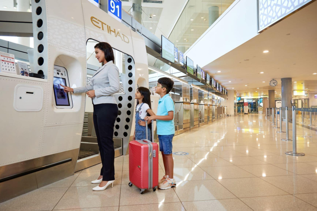 A woman uses an Etihad self-service check-in kiosk at the airport while two children wait with a suitcase