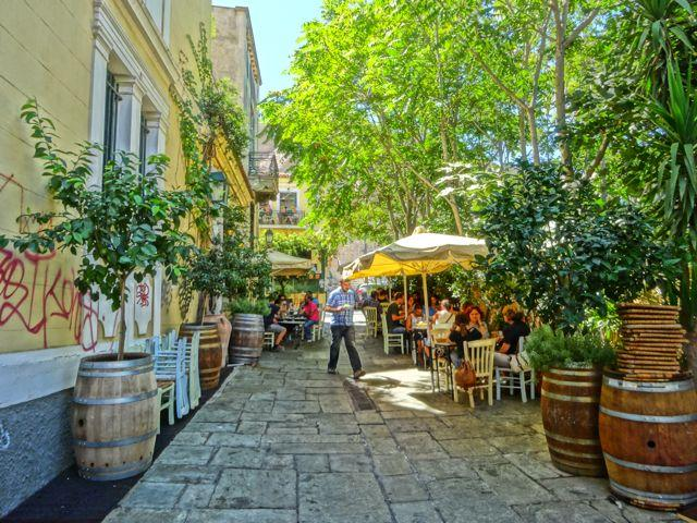 People dining at outdoor café tables on a narrow, tree-shaded paved street in Athens