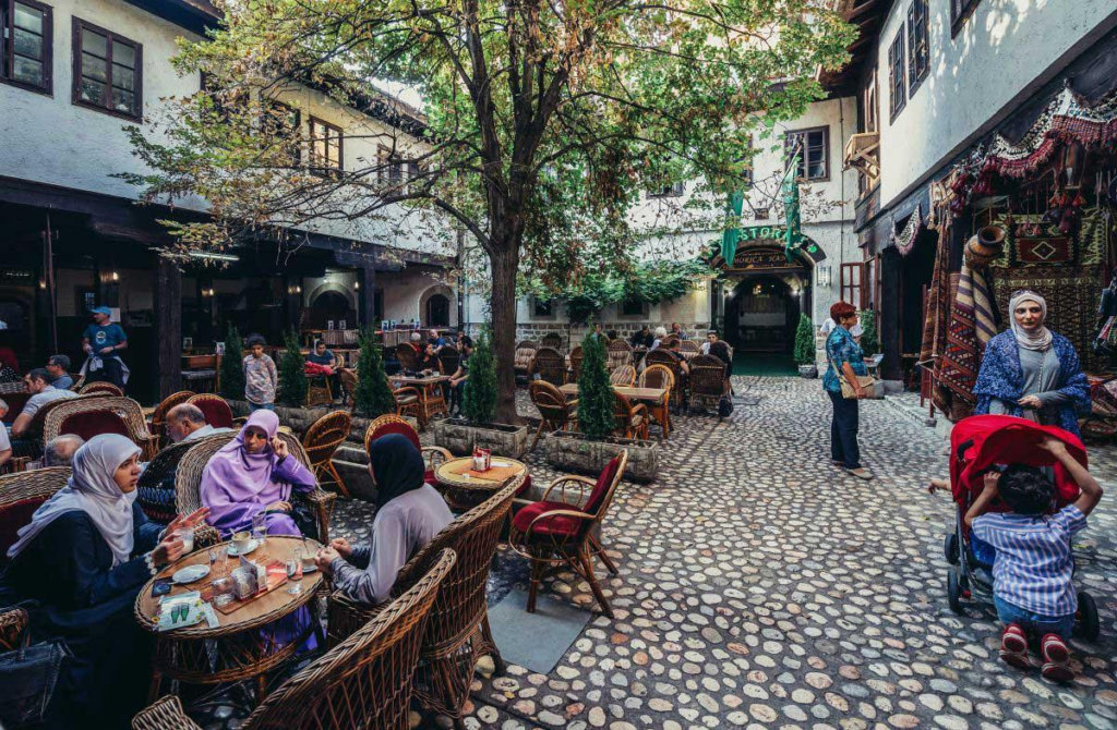 People sitting at wicker tables in the cobblestone courtyard of Morića Han, a traditional café in Sarajevo