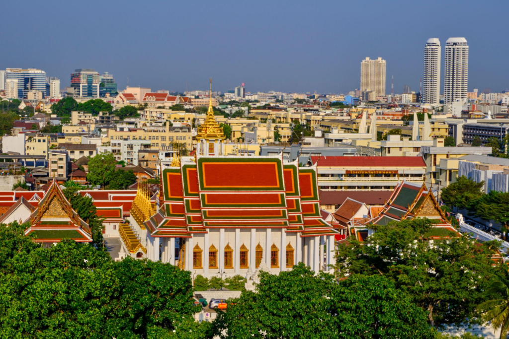 Traditional Thai temple with tiered red roofs and golden spires set against the modern Bangkok city skyline