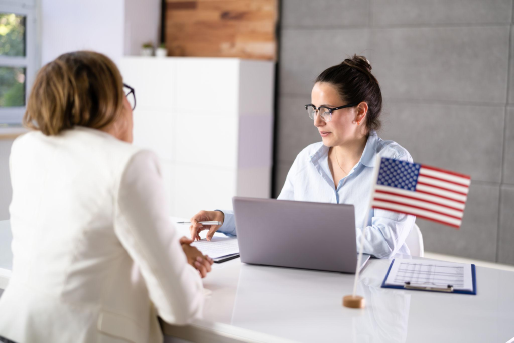 A woman conducting a U.S. visa interview with an applicant at a desk featuring a small American flag