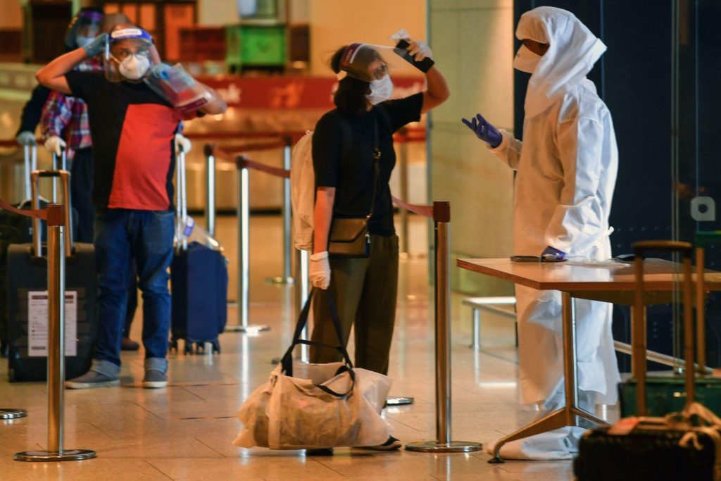 Travelers wearing masks and face shields at an airport health screening conducted by a worker in a hazmat suit