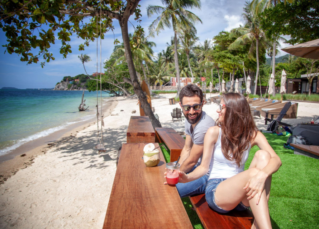 A couple smiles while enjoying drinks at a tropical beachfront bar with palm trees and clear blue water