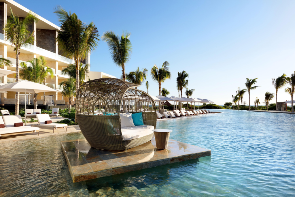 A modern resort pool area with an infinity edge, palm trees, and a wicker daybed on a platform in the water