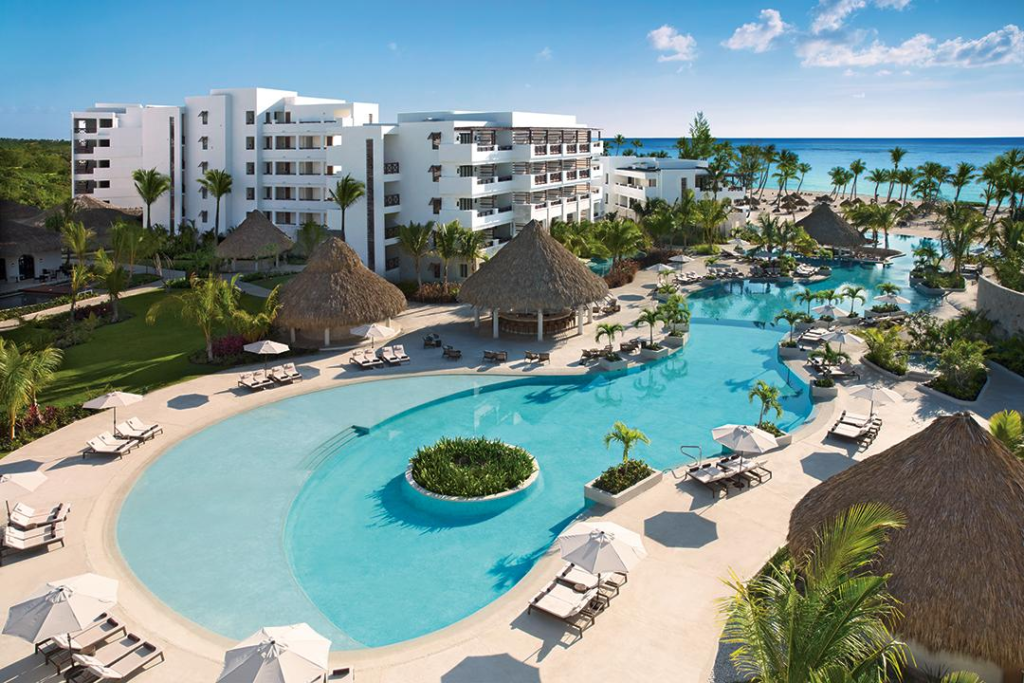 Modern white resort buildings overlooking a large, winding swimming pool area with thatched-roof bars and the ocean