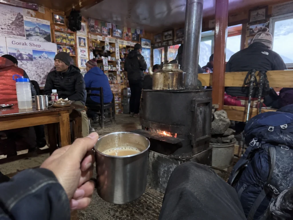 Hand holding a metal mug inside a cozy teahouse in Gorak Shep, with a central stove heating the room